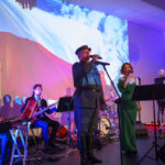 Grupa muzyków występuje na scenie z projekcją polskiej flagi w tle. Na pierwszym planie śpiewają mężczyzna i kobieta. Kolorowy wystrój podkreśla uroczystość wydarzenia.