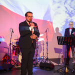 Mężczyzna w garniturze przemawia do mikrofonu na tle polskiej flagi. W tle perkusja, drugi mężczyzna stoi obok. Kolory czerwony, biały, niebieski.