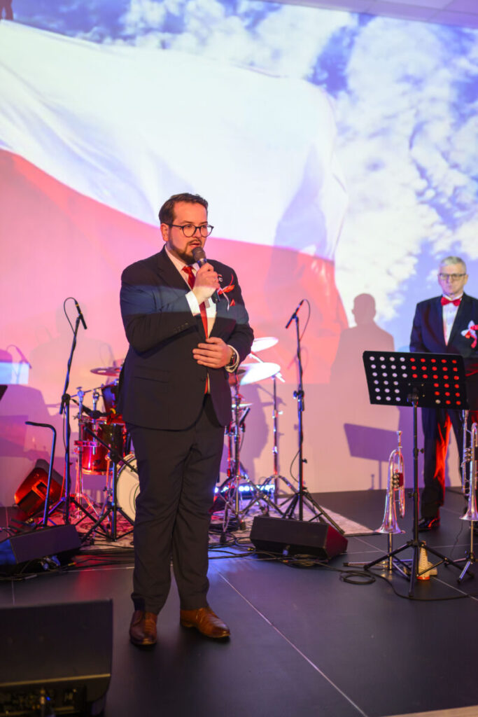 Mężczyzna w garniturze przemawia na scenie z mikrofonem. W tle biało-czerwona flaga i zestaw perkusyjny. Oświetlenie w ciepłych barwach.