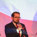 Mężczyzna w garniturze, trzyma mikrofon na tle flagi Polski. Dominują kolory czerwony i biały. W tle mikrofon na statywie.