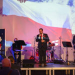 Na scenie stoi mężczyzna z mikrofonem, w tle flaga Polski. Obok muzycy z instrumentami, tło z wyświetloną flagą Polski. Dominuje czerwień i biel, sala pełna widzów.