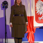 Kobieta w brązowym mundurze wojskowym stoi na tle flagi Polski z godłem. Oświetlenie sceniczne w tle. Po lewej lampa sceniczna.