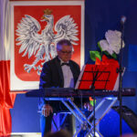 Mężczyzna w smokingu gra na klawiszach na tle flagi z orłem w koronie. Obok kwiatu białego i czerwonego. Niebieskie tło sceny.