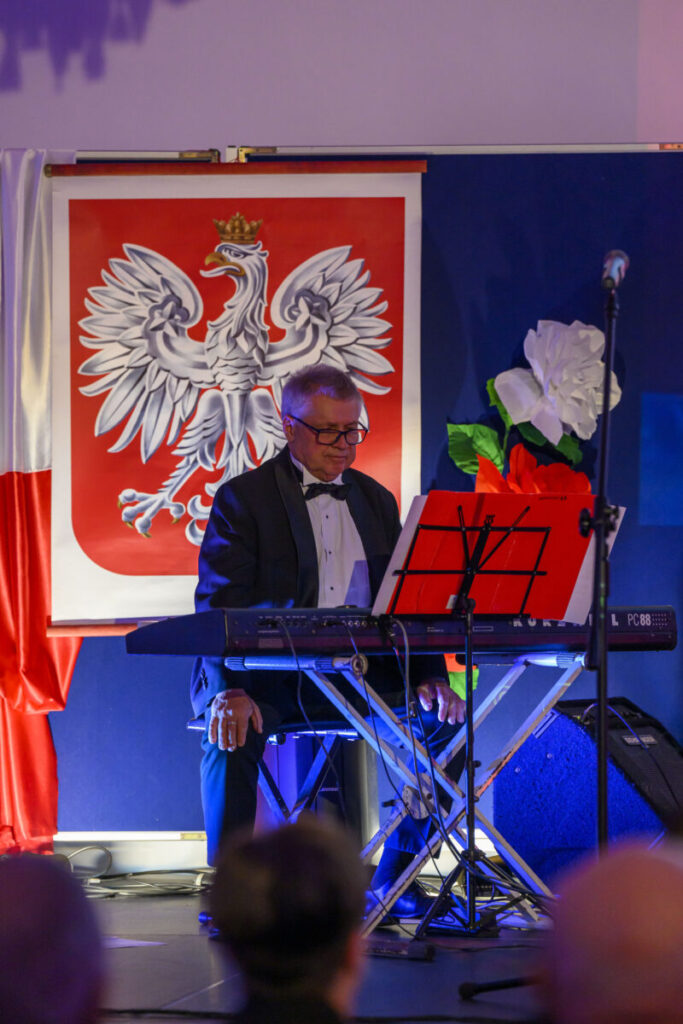 Mężczyzna w smokingu gra na klawiszach na tle flagi z orłem w koronie. Obok kwiatu białego i czerwonego. Niebieskie tło sceny.