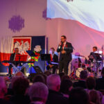 Na scenie występują muzycy i wokalista, tło z polską flagą i orłem, dominują kolory biały i czerwony.