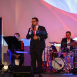 Mężczyzna w eleganckim garniturze śpiewa na scenie. Towarzyszą mu muzycy przy instrumentach na tle polskiej flagi. Światła w kolorze czerwonym i niebieskim.