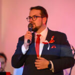 Na zdjęciu mężczyzna w okularach i garniturze z czerwonym krawatem trzyma mikrofon. Tło w ciepłych odcieniach różu. Po lewej niewyraźna postać.