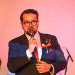 Nie wiem, kto jest na zdjęciu. Mężczyzna w garniturze trzyma mikrofon, ma brodę i okulary. Tło jest ciepłe, różowe z cieniami.