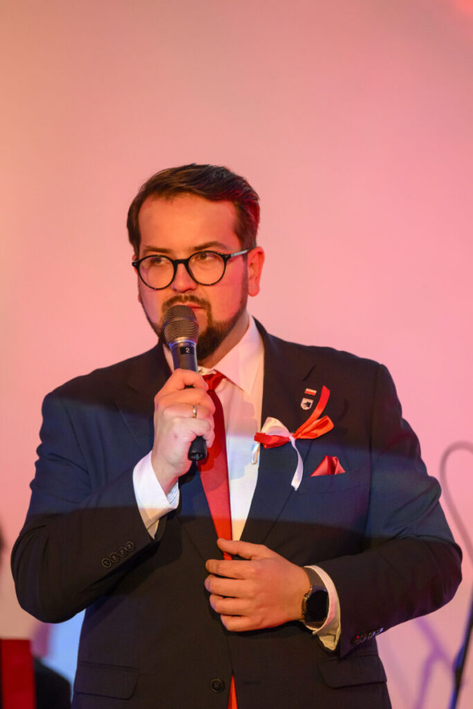 Mężczyzna w okularach mówi do mikrofonu, ubrany w ciemny garnitur z czerwonym krawatem. Tło jest jasnoróżowe.