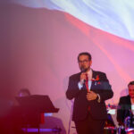 Mężczyzna w garniturze śpiewa na scenie z mikrofonem. Tło w kolorach czerwonym i białym, z muzykiem grającym na perkusji.