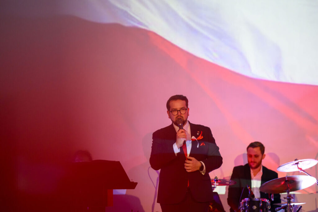 Mężczyzna w garniturze śpiewa na tle biało-czerwonej flagi. Ciepłe oświetlenie dominuje w kadrze. Obok perkusista.