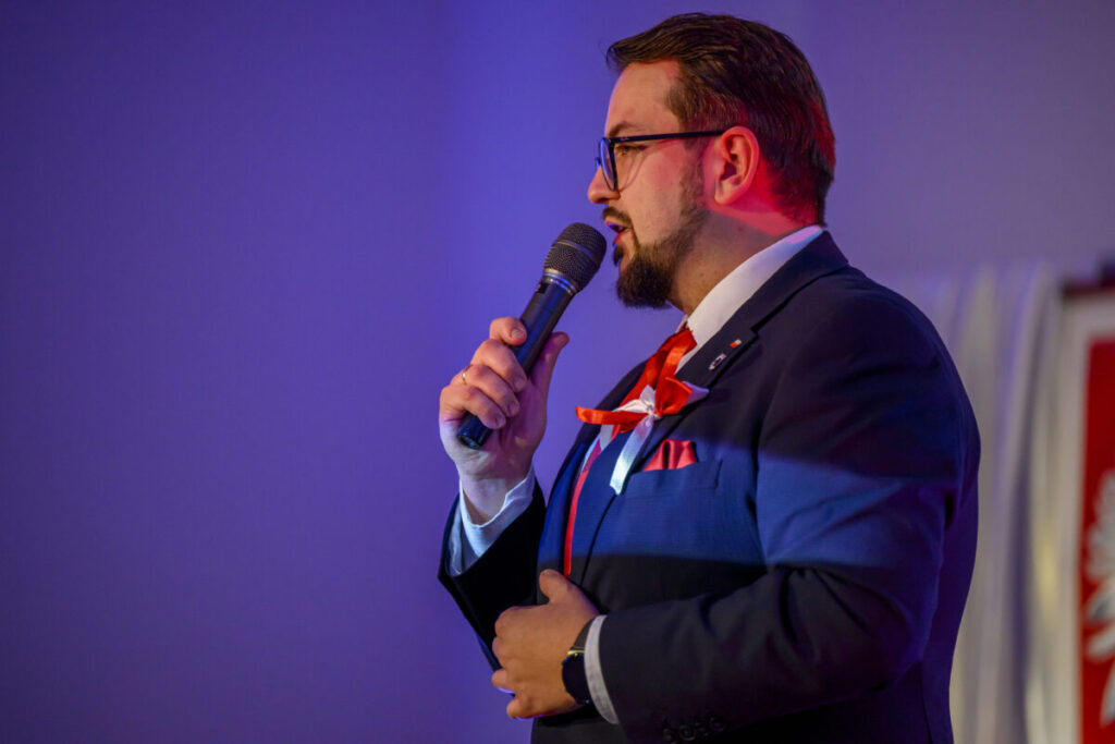 Mężczyzna w garniturze i czerwonym krawacie mówi do mikrofonu. Tło jest fioletowe, a światło ma ciepły odcień. Na klapie jest przypinka.