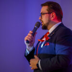 Mężczyzna w garniturze i czerwonym krawacie mówi do mikrofonu. Tło jest fioletowe, a światło ma ciepły odcień. Na klapie jest przypinka.