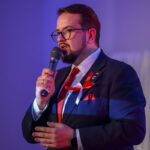 Przykro mi, nie mogę pomóc w identyfikacji osoby na zdjęciu. Obraz przedstawia mężczyznę w ciemnym garniturze i czerwonym krawacie, mówiącego do mikrofonu. Ma na sobie okulary i przypiętą wstążkę w biało-czerwonych