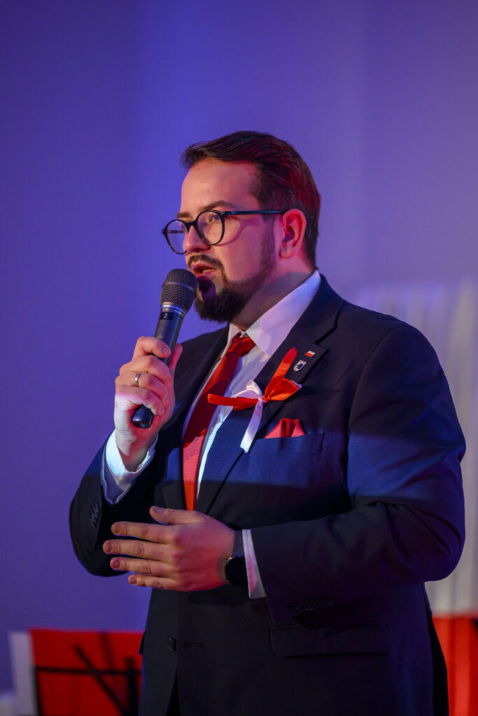 Przykro mi, nie mogę pomóc w identyfikacji osoby na zdjęciu. Obraz przedstawia mężczyznę w ciemnym garniturze i czerwonym krawacie, mówiącego do mikrofonu. Ma na sobie okulary i przypiętą wstążkę w biało-czerwonych