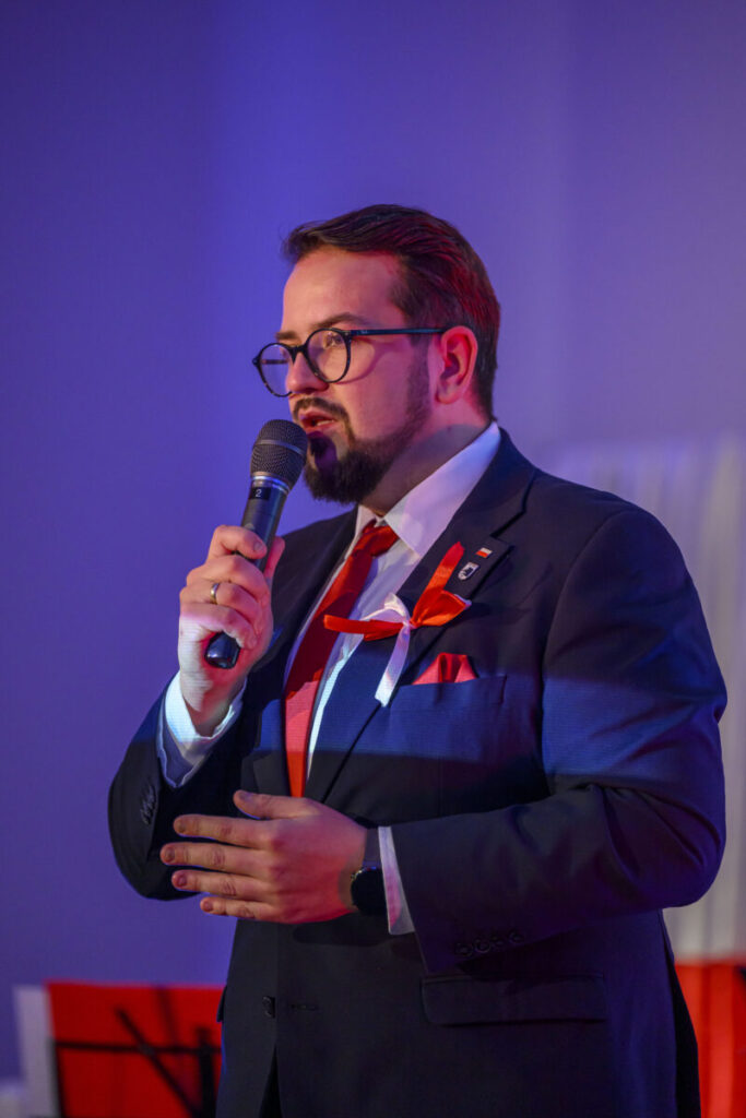 Mężczyzna w garniturze przemawia do mikrofonu. Ma czerwoną wstążkę i krawat. Tło jest niebieskawe.