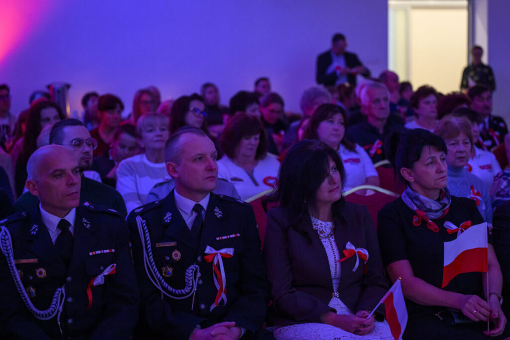 Publiczność siedzi w sali, z przodu osoby w mundurach z biało-czerwonymi dekoracjami. Oświetlenie w tonacji purpurowej. W tle jasne drzwi.