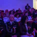 Publiczność siedzi w sali, z przodu osoby w mundurach z biało-czerwonymi dekoracjami. Oświetlenie w tonacji purpurowej. W tle jasne drzwi.