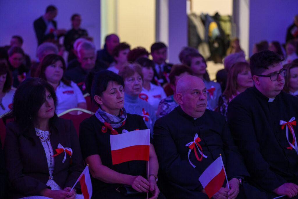 Sala wypełniona ludźmi trzymającymi polskie flagi. Osoby siedzą na czerwonych krzesłach, światło fioletowe. Tło to jasno oświetlone drzwi.