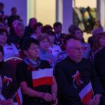 Sala wypełniona ludźmi trzymającymi polskie flagi. Osoby siedzą na czerwonych krzesłach, światło fioletowe. Tło to jasno oświetlone drzwi.