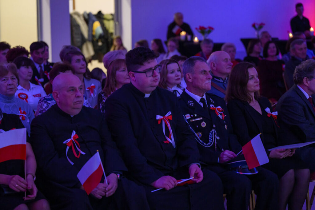 Grupa osób siedzi na widowni z biało-czerwonymi flagami, ubrana w eleganckie stroje. Atmosfera jest uroczysta z niebieskim oświetleniem. W tle widać kwiaty i świece.