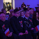 Grupa osób siedzi na widowni z biało-czerwonymi flagami, ubrana w eleganckie stroje. Atmosfera jest uroczysta z niebieskim oświetleniem. W tle widać kwiaty i świece.