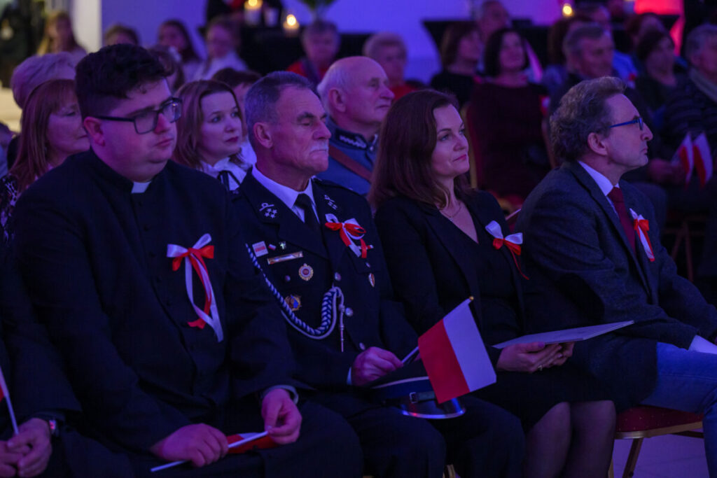 Na zdjęciu widać ludzi siedzących na wydarzeniu patriotycznym, ubranych elegancko z biało-czerwonymi kotylionami. Osoby trzymają polskie flagi. Tło jest ciemne, z fioletowym oświetleniem.