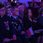 Na zdjęciu widać ludzi siedzących na wydarzeniu patriotycznym, ubranych elegancko z biało-czerwonymi kotylionami. Osoby trzymają polskie flagi. Tło jest ciemne, z fioletowym oświetleniem.