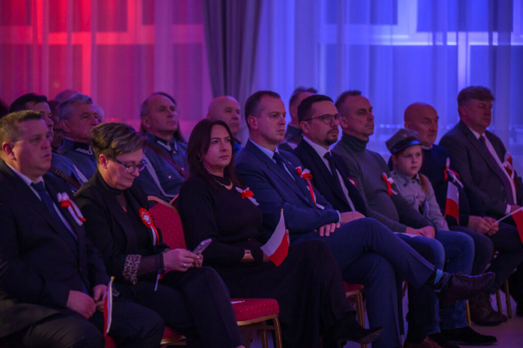 Grupa ludzi siedzących w sali, oświetlonej na czerwono i niebiesko. Wszystkie osoby mają biało-czerwone kokardy. Uczestnicy trzymają małe flagi Polski.