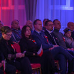 Grupa ludzi siedzących w sali, oświetlonej na czerwono i niebiesko. Wszystkie osoby mają biało-czerwone kokardy. Uczestnicy trzymają małe flagi Polski.