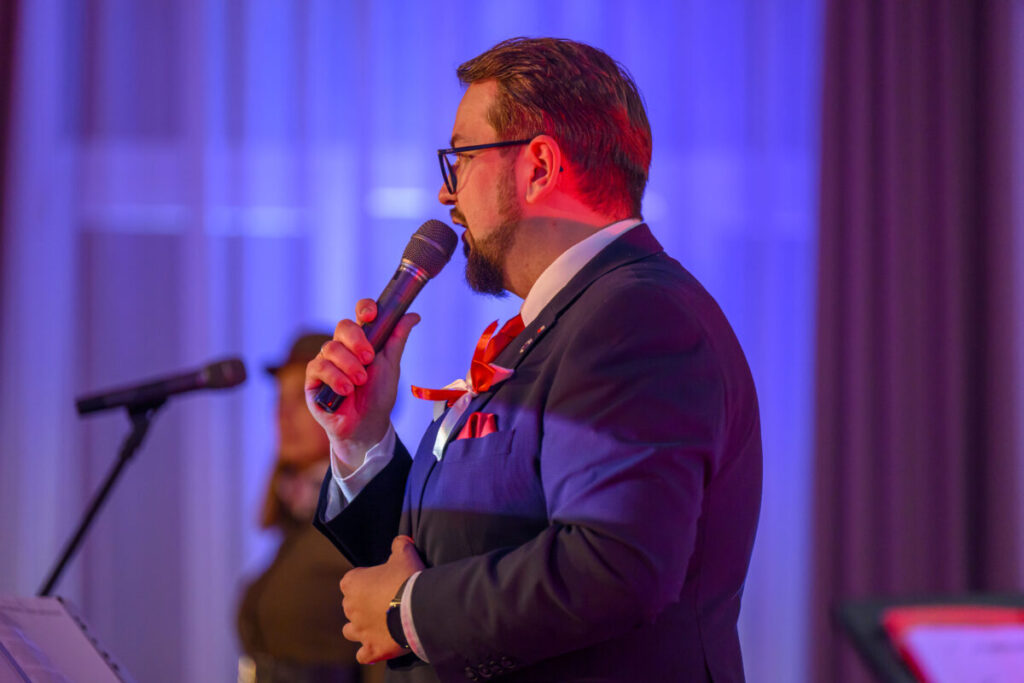 Mężczyzna w garniturze z czerwonym krawatem przemawia do mikrofonu. Tło w odcieniach niebiesko-fioletowych, widoczny drugi mikrofon. Scena oświetlona kolorowym światłem.