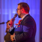 Mężczyzna w garniturze z czerwonym krawatem przemawia do mikrofonu. Tło w odcieniach niebiesko-fioletowych, widoczny drugi mikrofon. Scena oświetlona kolorowym światłem.