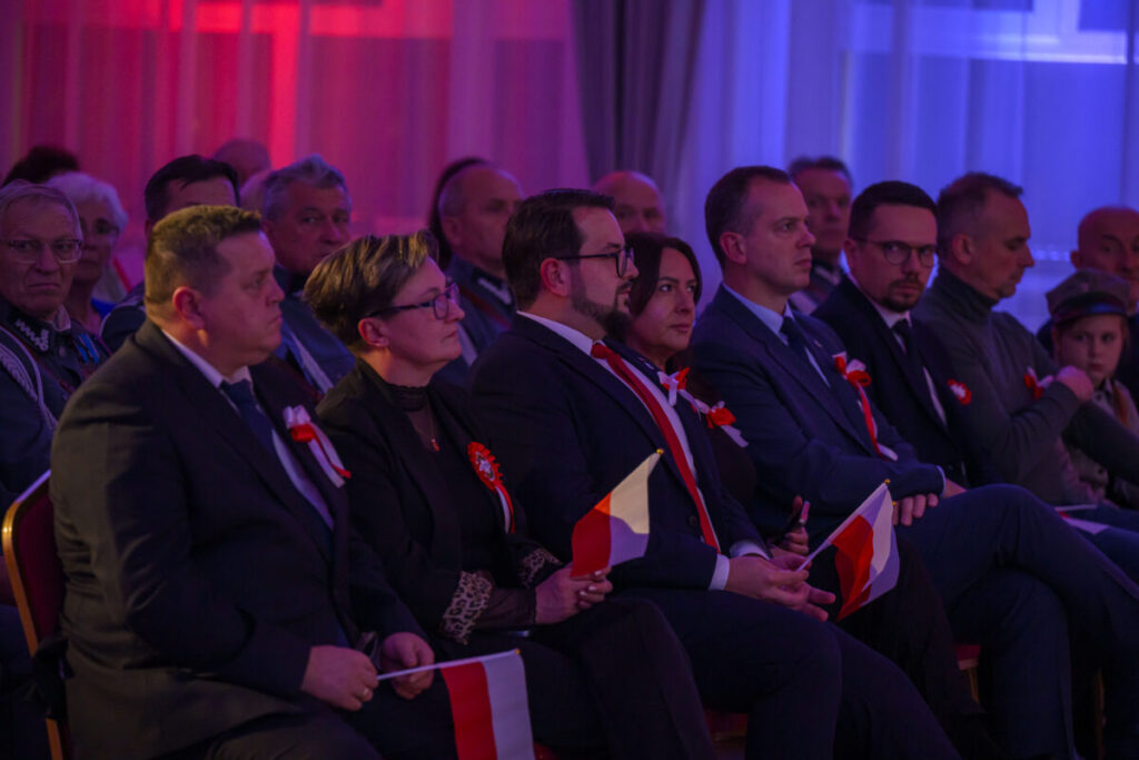 Grupa ludzi siedzących w ciemnym pomieszczeniu z czerwonym i niebieskim oświetleniem. Osoby mają flagi Polski i biało-czerwone kotyliony. W tle są ciemne zasłony.