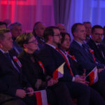 Grupa ludzi siedzących w ciemnym pomieszczeniu z czerwonym i niebieskim oświetleniem. Osoby mają flagi Polski i biało-czerwone kotyliony. W tle są ciemne zasłony.