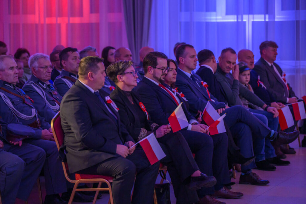 Grupa ludzi siedzi w sali, trzymając polskie flagi. Tło oświetlone jest na czerwono i niebiesko. Osoby ubrane są elegancko.