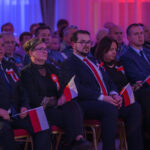 Grupa osób siedzi na krzesłach, trzymając polskie flagi. Wszyscy są elegancko ubrani, większość ma biało-czerwone kotyliony. Tło jest w niebiesko-czerwonych odcieniach.