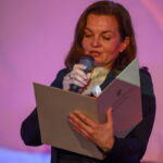 Kobieta trzyma mikrofon i czyta dokument na tle różowego światła. Elegancki strój, wyraziste oświetlenie.