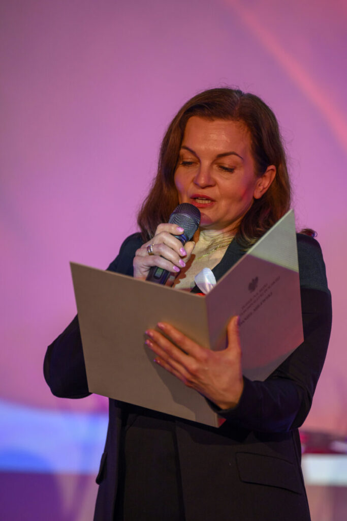 Kobieta trzyma mikrofon i czyta dokument na tle różowego światła. Elegancki strój, wyraziste oświetlenie.