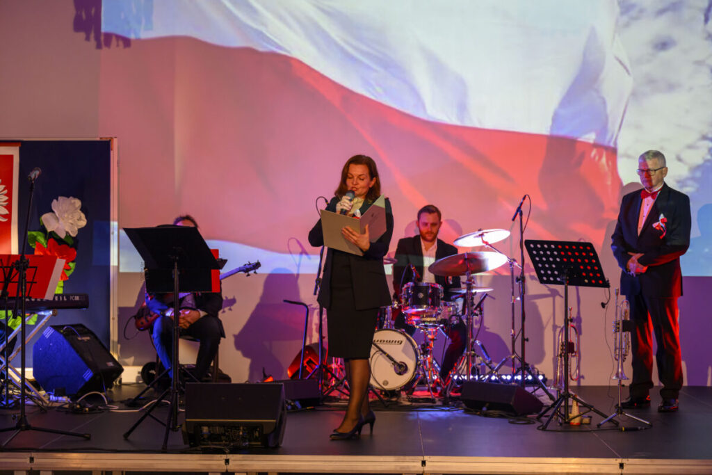 Na scenie kobieta przemawia do mikrofonu, w tle flaga Polski. Obok zespół muzyczny, dominują czerwone i białe kolory. Tło ma patriotyczny charakter.