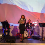 Na scenie kobieta przemawia do mikrofonu, w tle flaga Polski. Obok zespół muzyczny, dominują czerwone i białe kolory. Tło ma patriotyczny charakter.