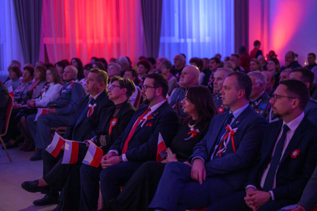 Grupa ludzi siedzi na sali, ubranych elegancko, z biało-czerwonymi akcentami. Światło jest niebiesko-czerwone. Tło to zasłony i ściany w kolorach eventu.