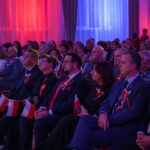 Grupa ludzi siedzi na sali, ubranych elegancko, z biało-czerwonymi akcentami. Światło jest niebiesko-czerwone. Tło to zasłony i ściany w kolorach eventu.