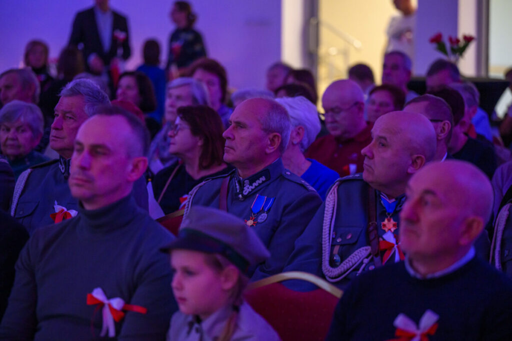 Grupa ludzi w mundurach siedzi na widowni. Dominują niebieskie i czerwone kolory. W tle rozmyte, jasne światło.