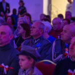 Grupa ludzi w mundurach siedzi na widowni. Dominują niebieskie i czerwone kolory. W tle rozmyte, jasne światło.