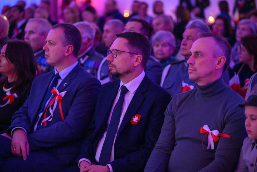 Grupa ludzi siedzi na uroczystości, ozdobiona biało-czerwonymi kokardkami. Ubrani w eleganckie stroje. Tło niebiesko-różowe.