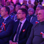 Grupa ludzi siedzi na uroczystości, ozdobiona biało-czerwonymi kokardkami. Ubrani w eleganckie stroje. Tło niebiesko-różowe.