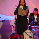 Kobieta w czarnej sukni przemawia do mikrofonu, trzymając kartki. W tle widać perkusistę i kolorowe oświetlenie sceniczne.