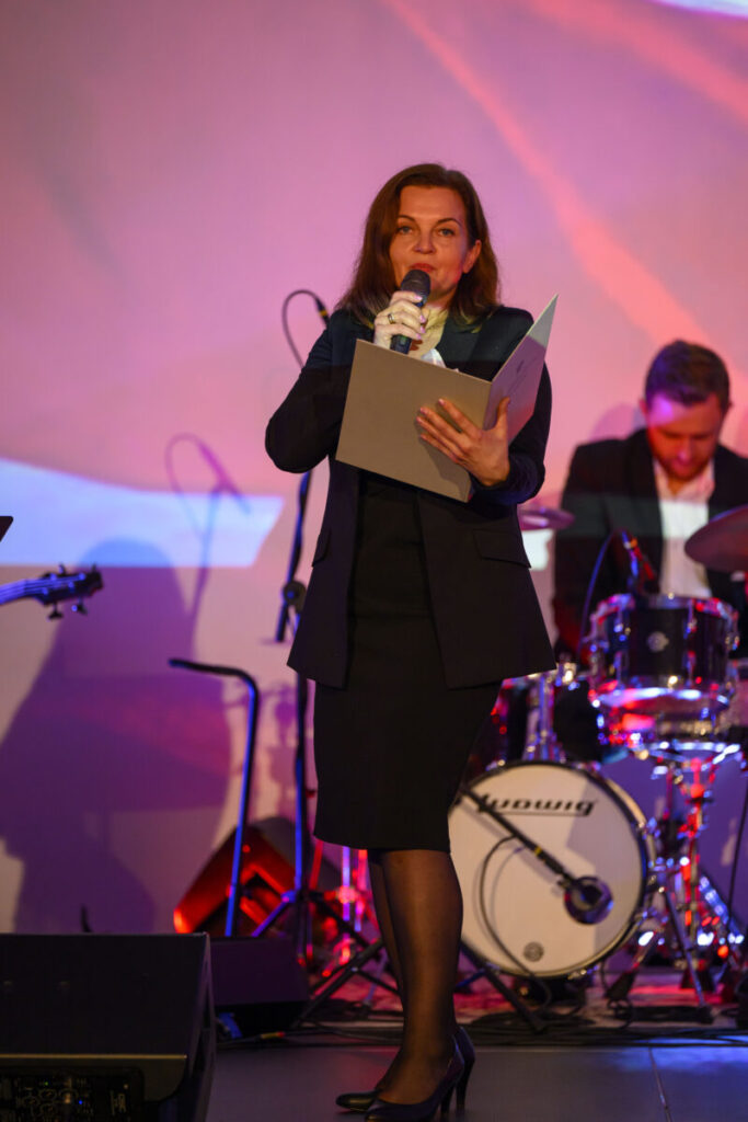 Kobieta w czarnej sukni przemawia do mikrofonu, trzymając kartki. W tle widać perkusistę i kolorowe oświetlenie sceniczne.