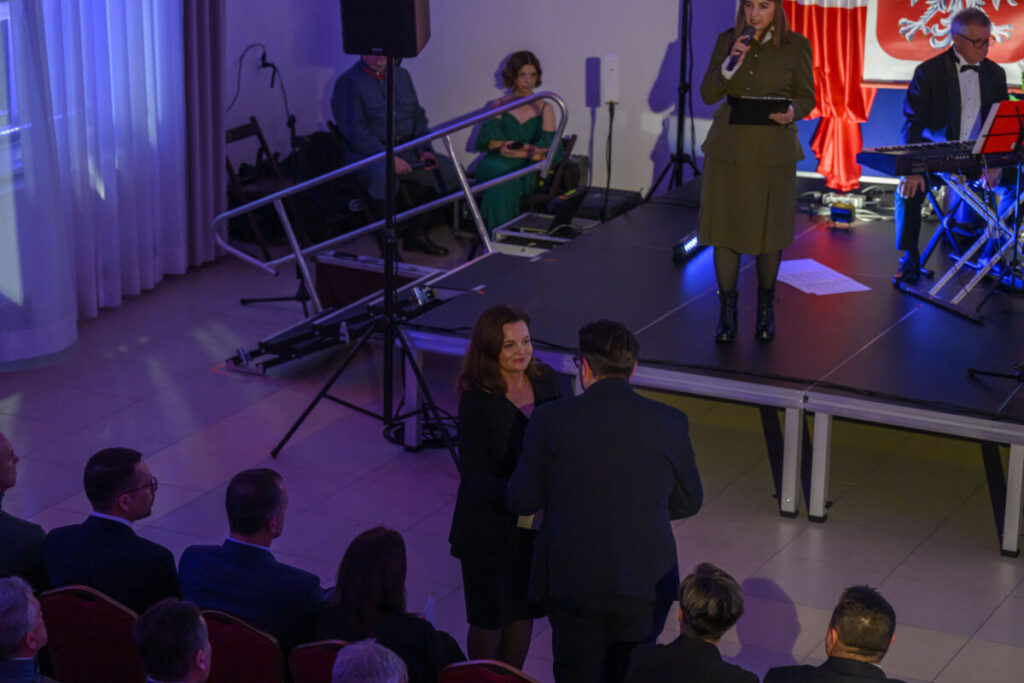 Pomieszczenie z ludźmi na uroczystości. Kobieta na scenie w khaki przemawia, obok muzycy. Dominują kolory czerwony i niebieski.