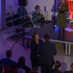 Pomieszczenie z ludźmi na uroczystości. Kobieta na scenie w khaki przemawia, obok muzycy. Dominują kolory czerwony i niebieski.